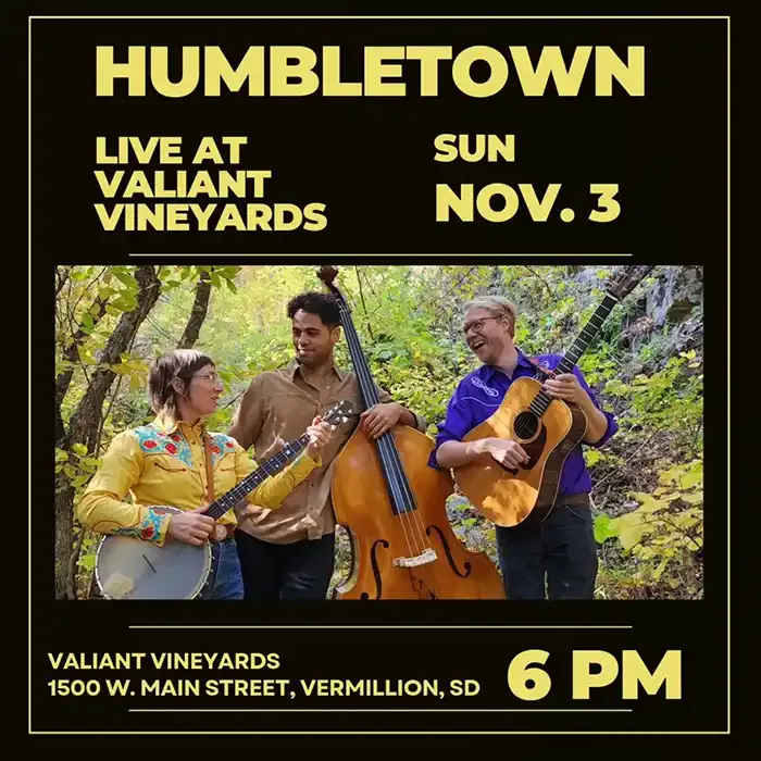 Promotional poster for a Humbletown concert at Valiant Vineyards on Sunday, November 3 at 6 PM. The poster features a photo of the three band members outdoors among fall foliage, smiling and playing instruments including a banjo, upright bass, and acoustic guitar. Text includes the venue address: 1500 W. Main Street, Vermillion, SD