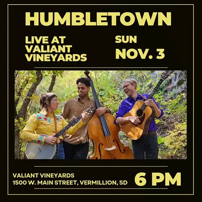 Promotional poster for a Humbletown concert at Valiant Vineyards on Sunday, November 3 at 6 PM. The poster features a photo of the three band members outdoors among fall foliage, smiling and playing instruments including a banjo, upright bass, and acoustic guitar. Text includes the venue address: 1500 W. Main Street, Vermillion, SD