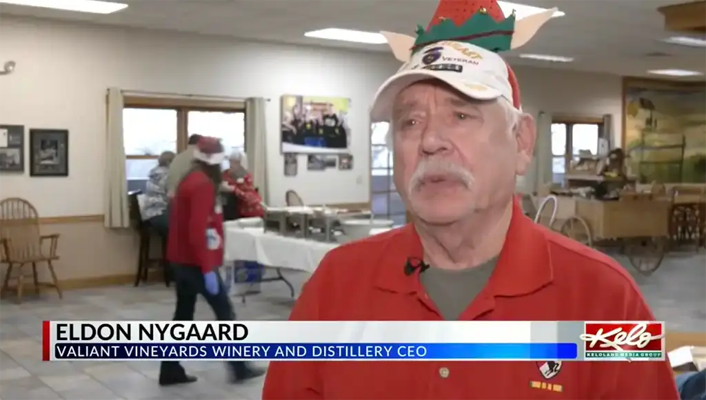 Eldon Nygaard, CEO of Valiant Vineyards Winery and Distillery, speaks during a news interview inside the winery. He is wearing a red shirt and a festive holiday hat, with people preparing food in the background and the KELOLAND news graphic displayed at the bottom of the screen.