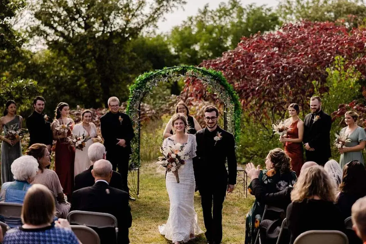 Newly married couple walking down the aisle beneath a green arch at an outdoor fall wedding, with wedding party and guests applauding.
