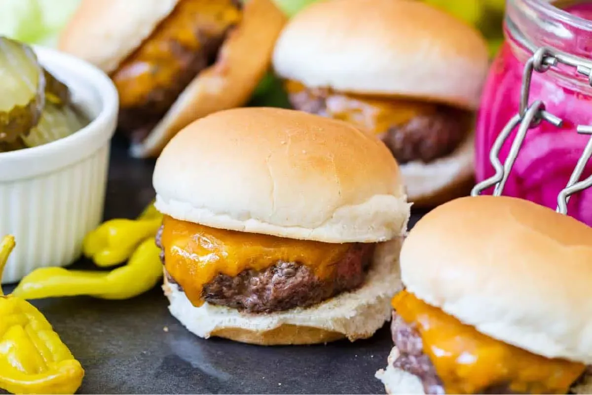Close-up of several cheeseburger sliders on soft buns, topped with melted cheddar and arranged on a platter with pickles, pickled onions, and pepperoncini peppers on the side.