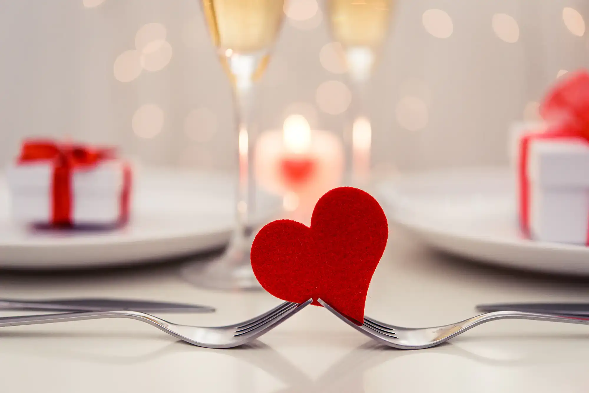 A romantic table setting with two intertwined forks holding a small red heart, with champagne glasses, a lit candle, and wrapped gifts softly blurred in the background.